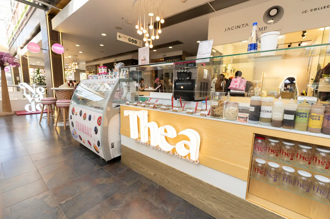 Cafe counter in a shopping mall with a glass-topped display of toppings and jars; Thea logo on the wooden front and a gelato freezer to the left.