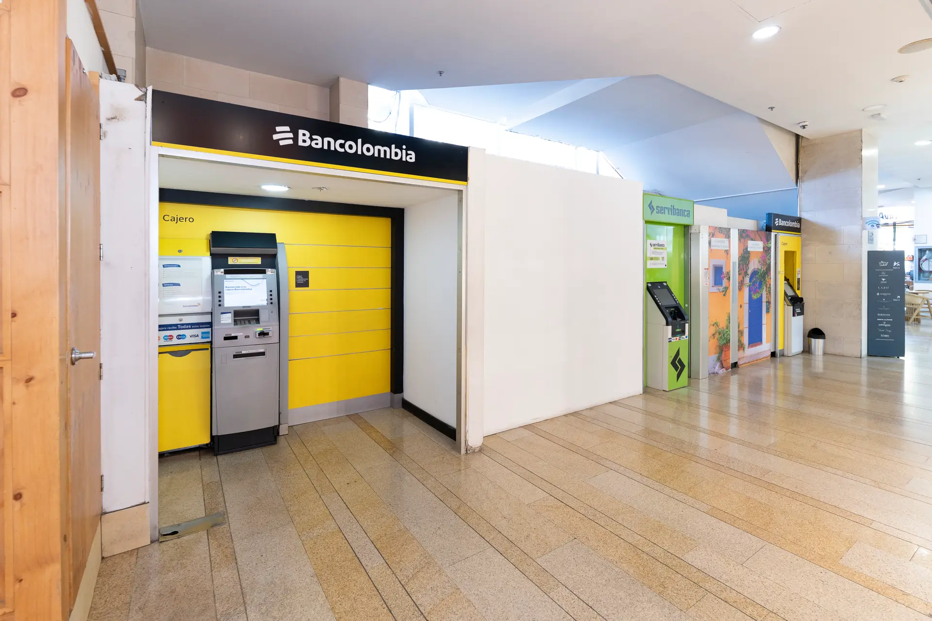 Row of bank ATMs and service kiosks inside a mall, featuring a bright yellow Banco Colombia ATM facade on the left and colorful machines along the corridor