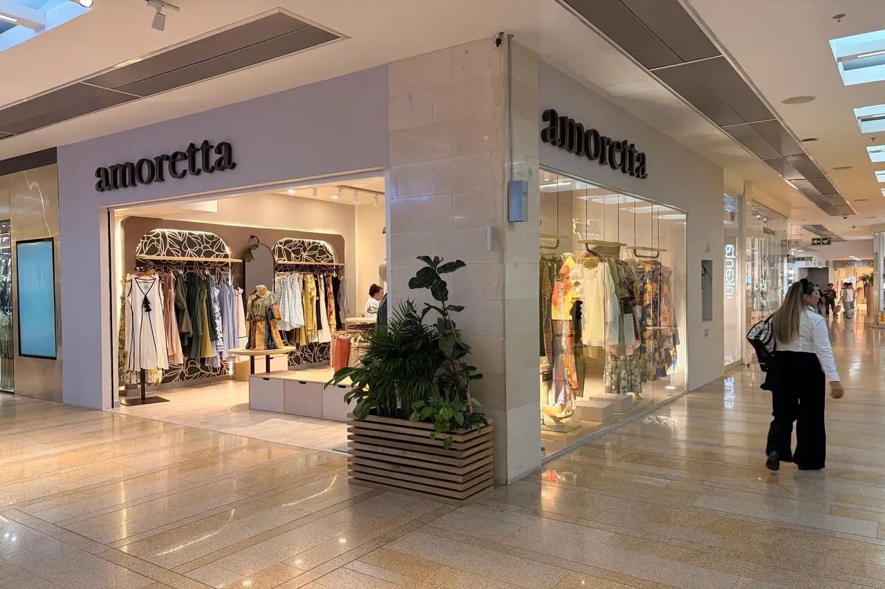 Clothing storefront named 'amoretta' with mannequins and racks of clothes inside, a planter near the entrance in a bright mall.