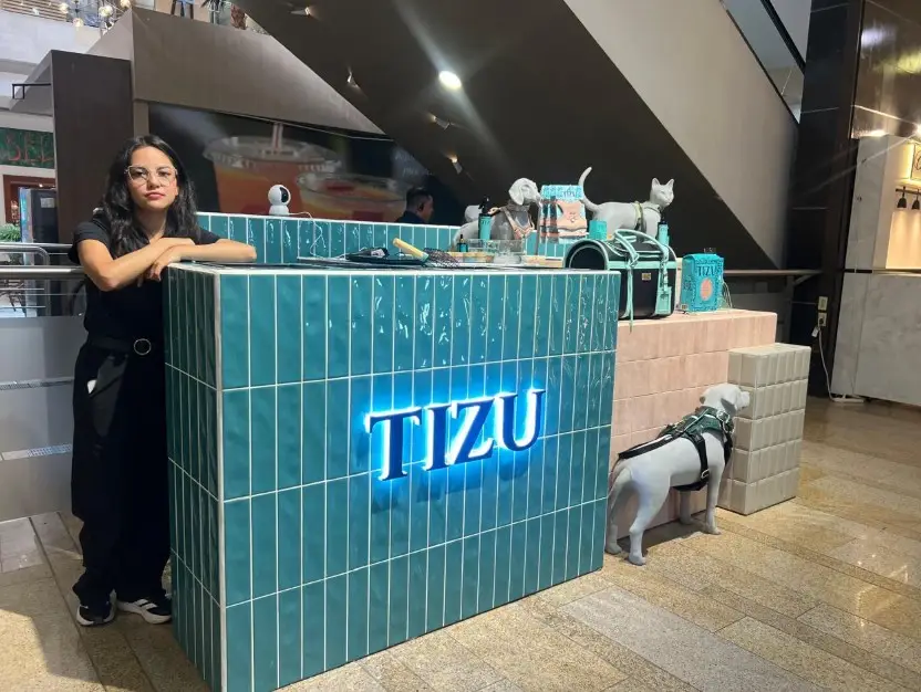 Woman leaning on a teal tiled counter with a glowing TZU sign; a dog statue and cat figurines are on display in a mall cafe area.